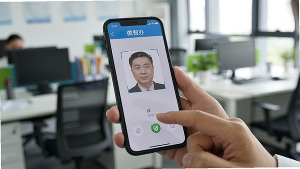 用户使用手机操作徽智办应用进行实名认证的场景，真人手部持手机，屏幕显示人脸识别框，背景为模糊的办公室环境，光线充足，突出手机屏幕内容，屏幕中需有