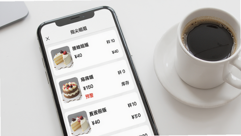 手机界面展示商品管理页面，列表显示多个商品条目，每个商品有缩略图、名称、价格和库存数字，其中一款蛋糕库存显示“预警”红色字体；背景为简约白色桌面，旁边放着一杯咖啡。整体干净、专业，体现指尖姐姐商家版智能库存功能。