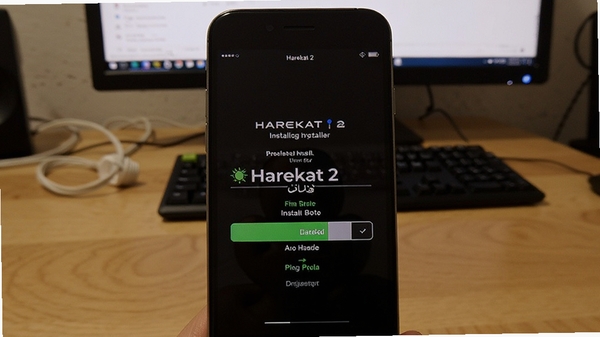 一张展示Harekat 2安装器在手机上运行的实拍照片,手机屏幕中显示安装进度条,背景为木桌与电脑,营造真实使用场景