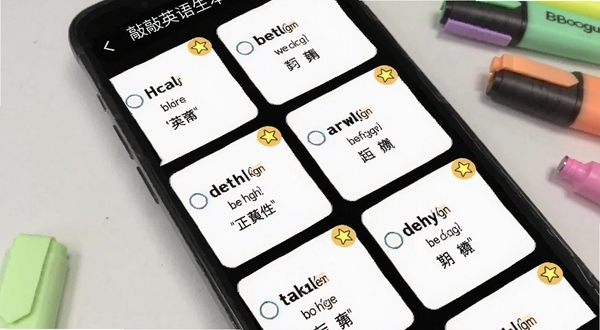 画面呈现敲敲英语生词本界面，手机屏幕中列出一张张单词卡片，每张卡片上有英文单词、音标和中文释义，部分卡片带有星标收藏图标，背景是简约的浅灰色桌面，旁边散落着几支彩色荧光笔，整体风格干净利落，适配Boogu模型生成