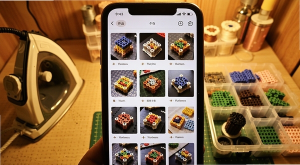 拼豆派个人作品展示页面,手机屏幕内呈现用户上传的拼豆成品照片墙,每张作品配有名称标签,画面主体为手机界面特写,背景为暖色调手工工作室,桌面摆放熨斗与拼豆收纳盒,整体氛围温馨
