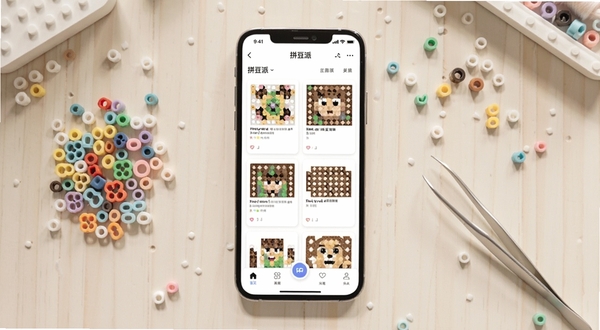 拼豆派软件主界面全貌展示,画面中央是手机屏幕正视图,屏幕内呈现拼豆派的应用首页,包含拼豆图纸卡片列表与底部导航栏,背景为柔和的浅色手工桌面,周边点缀彩色拼豆颗粒与镊子工具,整体风格清新明亮,适配Boogu模型生成