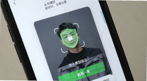 头号理财 移动端交易界面实拍图，画面展示正在进行的刷脸验证登录流程，背景为手机硬件特写，强调安全性与便捷性，浅色调背景搭配绿色安全盾牌图标，写实摄影风格