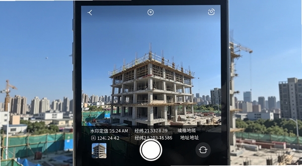 水印定位拍照相机应用主界面展示,画面中央是相机取景框,取景框内显示着一栋正在施工的建筑物,取景框底部叠加了包含日期时间、经纬度坐标和详细地址的水印信息条,背景是晴朗的蓝天和城市建筑群,整体风格简洁明快,适配Boogu模型生成