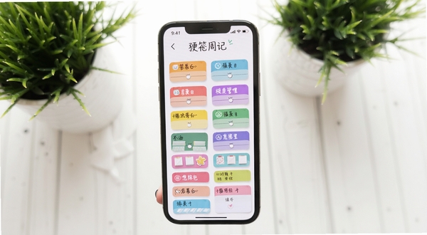 便签周记多彩标签分类管理界面,手机屏幕展示彩色标签卡片整齐排列,背景为白色桌面与绿植,色调柔和明亮,突出分类管理功能