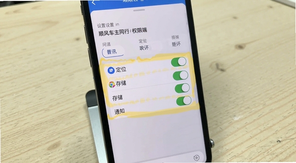 顺风车主同行手机端权限设置界面截图，画面主体为手机屏幕显示应用权限管理页面，高亮标注定位、存储、通知三个权限开关，背景为浅色木纹桌面与手机支架，整体风格清晰直观，突出操作引导