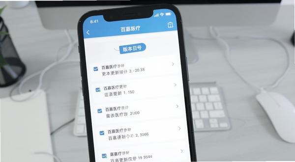百嘉医疗版本更新界面截图,画面主体为手机屏幕显示更新日志列表与版本号标签,背景为浅灰色办公桌面,风格简洁清晰