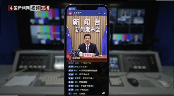 中国新闻网视频直播界面展示，画面中央为手机屏幕竖版特写，屏幕中正在播放新闻发布会直播画面，画面下方有实时评论滚动条，背景区域为演播室控制台虚化效果，突出直播的现场感与互动性