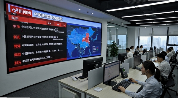 中国新闻网新闻编辑室工作场景，画面主体为一块巨大的新闻直播大屏，屏幕上显示中国新闻网实时新闻更新界面与地图定位标记，背景区域为现代化编辑办公区，多名编辑人员正在电脑前忙碌，整体氛围紧张有序，突出新闻时效性与权威性
