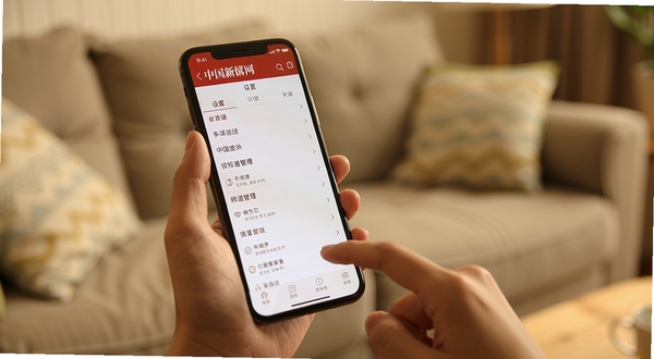 中国新闻网手机端设置界面与操作手势示意图，画面主体为一只手握持智能手机，屏幕上显示中国新闻网设置菜单与频道管理界面，背景区域为居家沙发环境虚化，整体色调温暖舒适，体现用户日常阅读新闻的轻松场景