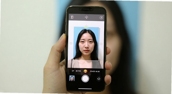 证件照拍摄院移动端操作界面截图，展示用户手持手机拍摄证件照的场景，屏幕中显示取景框与拍摄按钮，光线明亮，背景为纯色墙面
