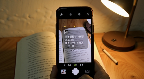 译语翻译通拍照翻译功能演示图,画面展示手机摄像头对准一本外文书籍页面,屏幕取景框中叠加显示识别出的文字与中文译文,背景为木质桌面与暖色台灯光线,整体氛围安静专注