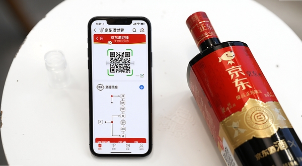 京东酒世界正品溯源功能展示图，画面中央是一部手机，屏幕显示京东酒世界APP的商品溯源页面，页面中有二维码扫描入口和溯源信息链条，手机旁边摆放着一瓶包装精美的白酒，背景为简洁的白色桌面，光线明亮清晰，突出正品保障主题