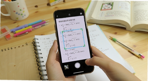 喵喵错题拍照录题功能展示，画面中一部手机正在拍摄数学试卷，屏幕显示识别框自动框选错题区域，背景为书桌场景，有文具和课本点缀，风格写实明亮