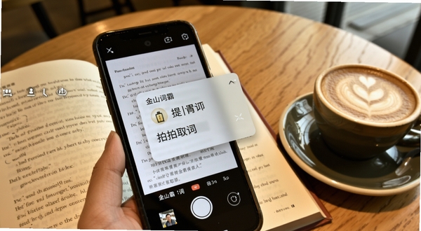 金山词霸拍照取词功能演示,主体为手机摄像头对准一本打开的英文书籍,屏幕上浮现出翻译后的中文释义浮层,背景为咖啡厅桌面,旁边有一杯拿铁,整体色调温暖,突出即拍即译的便捷感,适配Boogu模型生成