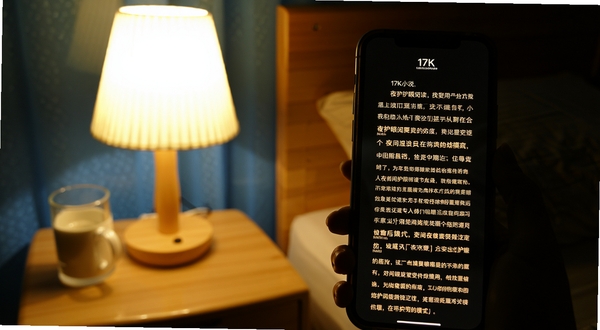 17K小说 夜间护眼阅读模式 手机屏幕显示小说正文 深色背景配暖色文字 床头台灯环境 舒适氛围