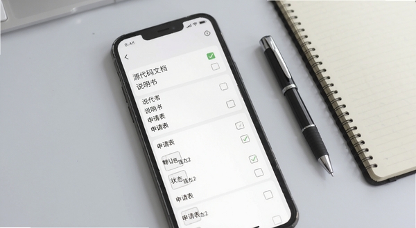 软件著作权应用内登记材料清单界面截图,手机屏幕显示分类清晰的材料列表,包含源代码文档、说明书、申请表等条目,每个条目右侧有状态勾选框,背景为浅灰色办公桌面环境,旁边放置一支笔和一本笔记本,体现办公场景,画面风格清爽整洁