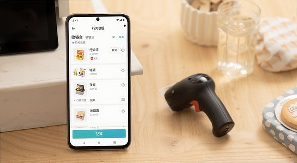 行知收银主界面展示,画面主体为一部竖屏安卓手机居中偏左位置,手机屏幕上呈现行知收银的收银台页面,可见商品列表与结算按钮;背景为浅色木质桌面,右侧区域摆放一台小型扫码枪和几件日用品,整体色调温暖明亮,写实产品摄影风格,细节清晰,适合下载站展示用图