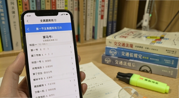 安驾课堂全真题库练习界面，手机屏幕显示科目一题目与解析内容，旁边有章节分类列表，背景为书房学习环境，画面中有交通法规书籍与荧光笔，光线明亮柔和
