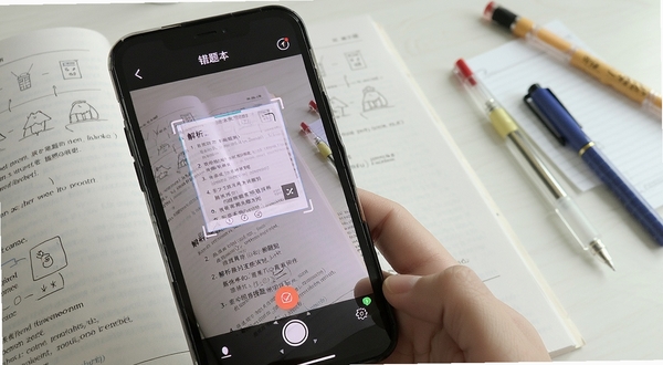 作业拍照搜题错题本拍照识别界面展示,手机对准练习册题目,屏幕出现识别框与解析步骤,背景为书桌与文具,画面清晰明亮