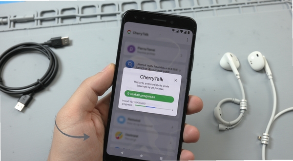 CherryTalk安装与权限设置操作场景图，画面中央为手持安卓手机的特写，屏幕显示权限授权弹窗与安装进度条，背景为简洁的浅灰工作台，旁边放置数据线与耳机等配件，整体风格写实清晰，突出操作指引感
