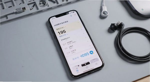 ADB助手版本更新界面展示,手机屏幕中呈现更新日志列表与版本号标识,背景为简洁的浅灰色工作台面,搭配数据线耳机等数码配件,整体色调清爽,适配Boogu模型生成