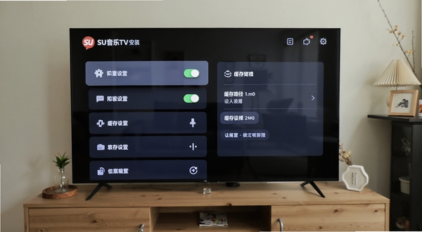 SU音乐TV安装设置界面，电视屏幕上显示权限开关与缓存路径选择面板，UI为深色卡片风格，画面主体为设置列表区域，背景为客厅电视柜场景